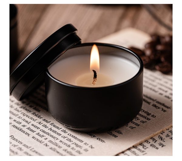 Black matte scented candle in a tin container with a burning flame, placed on an open book