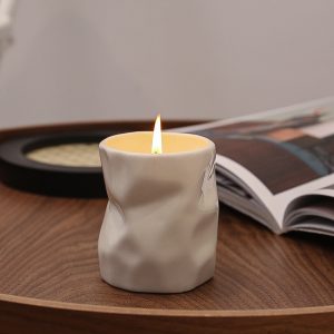 Modern crumpled white ceramic scented candle with a burning flame, placed on a wooden table next to an open book