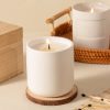 Lit white ceramic scented candle on a wooden coaster with a wicker tray in the background